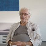 Halmer sitzt in Wartebereich mit gefaltenen Händen und schaut in Kamera