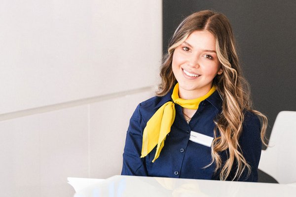 L&auml;chelnde junge Frau mit langem, gewelltem Haar, tr&auml;gt dunkelblaue Bluse und gelbes Halstuch, sitzt an einem Empfangstresen