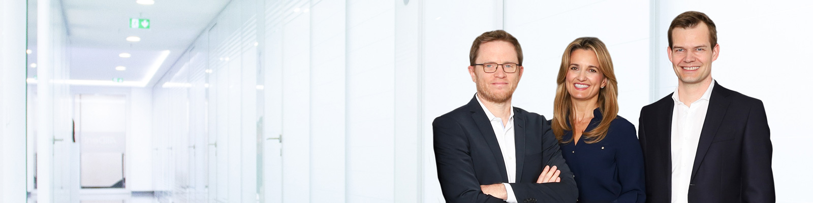 Drei Personen in Businesskleidung stehen in einem hellen Büroflur und lächeln in die Kamera