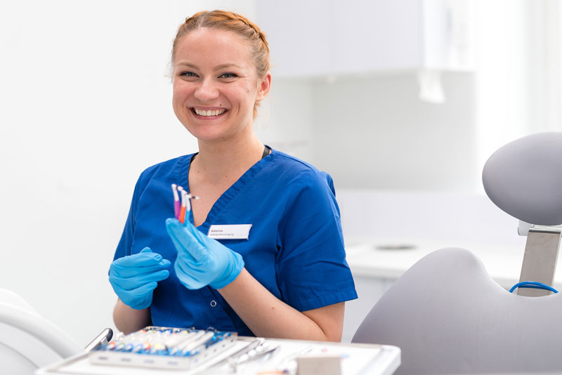 ZMP in blauer Arbeitskleidung und blauen Handschuhen hält Zahnfarbskala und lächelt in moderner Praxis