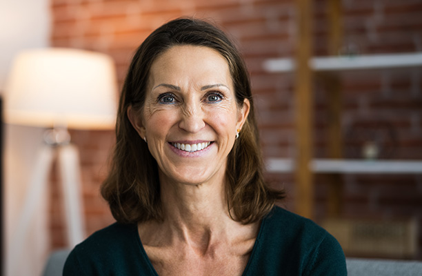 Frau mit braunen Haaren sitzt in Wohnzimmer und lächelt in Kamera