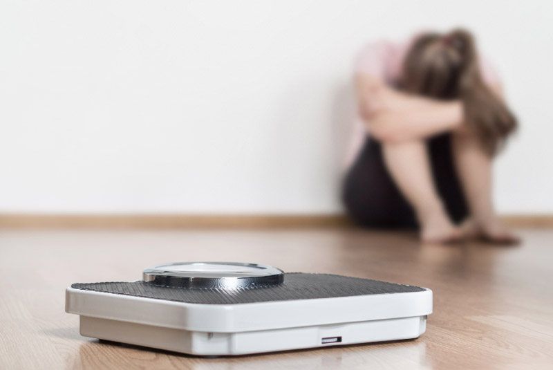 Personenwaage auf dem Fußboden, im Hintergrund lehnt eine Frau an der Hand und hat den Kopf in den Armen vergraben