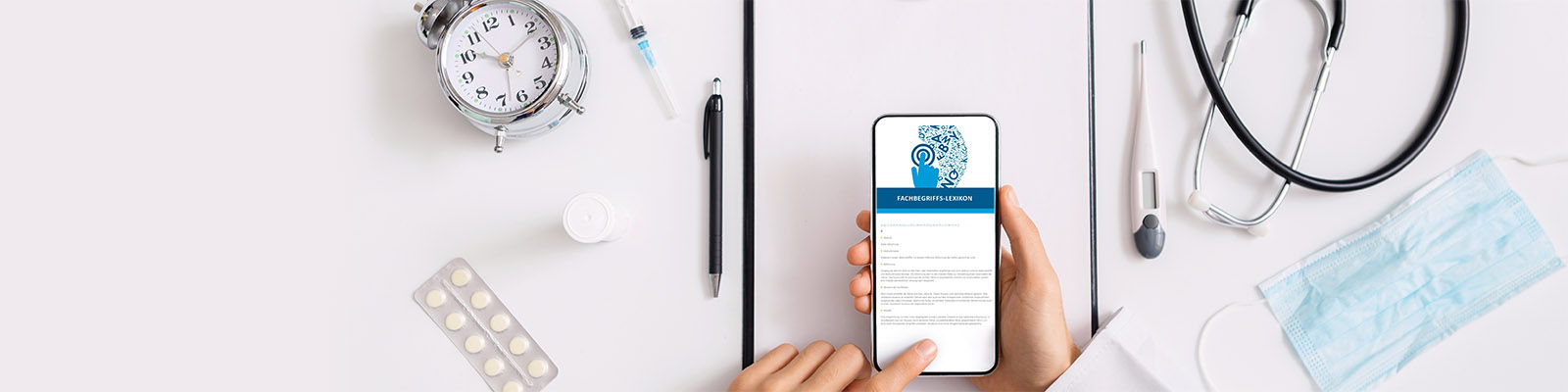 Finger tippt auf Handy mit aufgerufener Seite "Fachbegriffslexikon". Auf dem Tisch darunter liegt ein Stifr, Stetoskop, Tabletten, Mundschutz